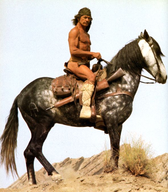 Charles Bronson habillé en indien, les cheveux longs et le torse nu, monté à cheval