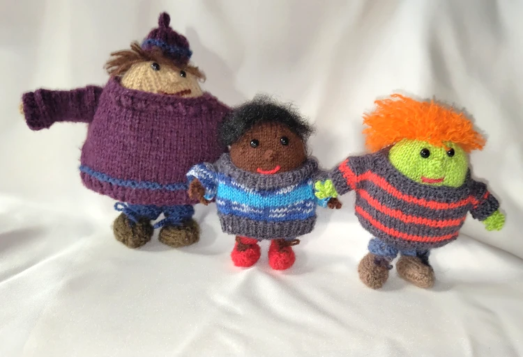 Trois personnages ovoïdes tricoté, le premier a le teint beige et un pull grenat, le second est chocolat avec un peu bleu et le troisième est roux et vert vif, son pull est gris rayé rouge.